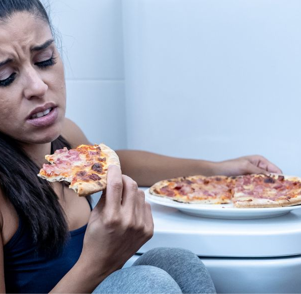 Kvinde sidder p[ badeværelses gulvet og spiser pizza ved siden af toilettet