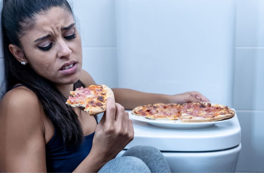 Kvinde sidder p[ badeværelses gulvet og spiser pizza ved siden af toilettet