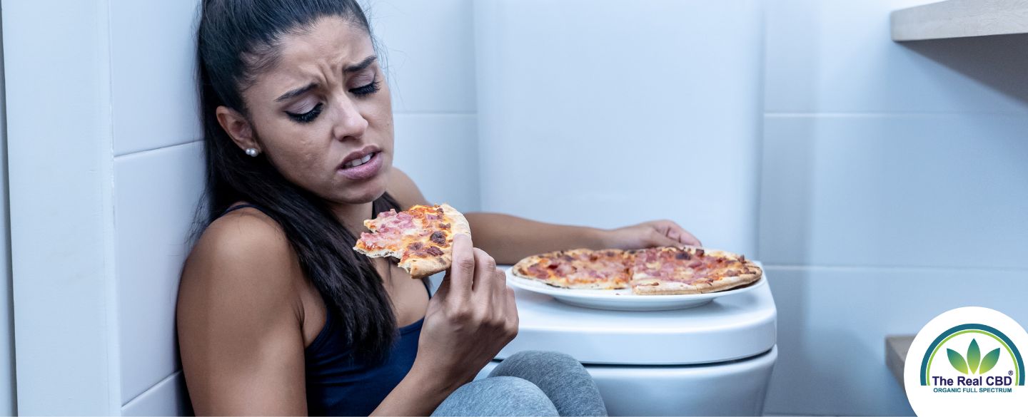 Kvinde sidder p[ badeværelses gulvet og spiser pizza ved siden af toilettet
