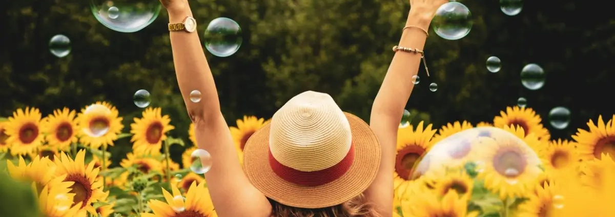 Mujer con sombrero de verano en un campo de girasoles, levantando los brazos entre burbujas en el aire
