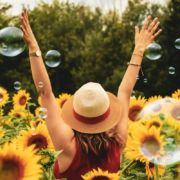 The-Real-CBD-Beneficios-de-aciete-de-CBD Mujer con sombrero de verano en un campo de girasoles, levantando los brazos entre burbujas en el aire