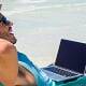 Hombre en una hamaca en la playa con su ordenador portátil trabajando bajo el sol.