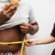 Vrouw houdt een meetlint rond de taille van een dikke man terwijl hij een hamburger eet