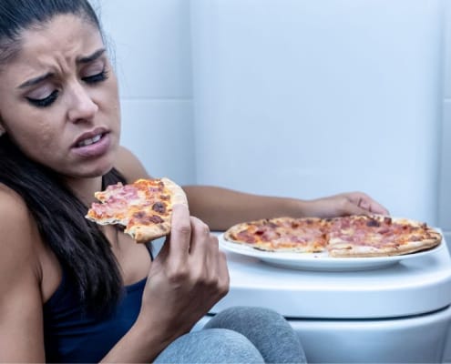 Frau isst Pizza auf dem Badezimmerboden neben der Toilette und sieht verzweifelt aus