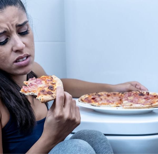 Vrouw eet pizza op de badkamervloer naast het toilet en kijkt bedroefd