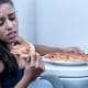 Vrouw eet pizza op de badkamervloer naast het toilet en kijkt bedroefd